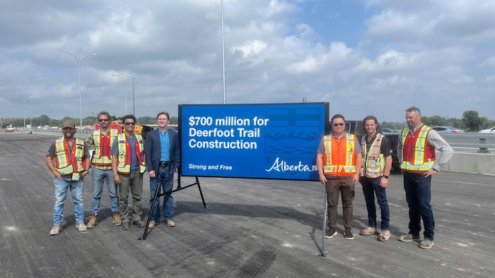 Devin Dreeshen et un groupe de six travailleurs de la construction portant des gilets de haute visibilité posent sur une route fraîchement asphaltée de part et d'autre d'une grande affiche bleue du gouvernement de l'Alberta annonçant 700 millions de dollars pour la construction de Deerfoot Trail.