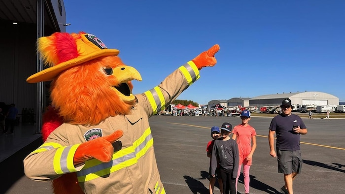 Une mascotte représentant un phénix vêtu d’un uniforme de pompier lève un bras dans les airs, dehors.
