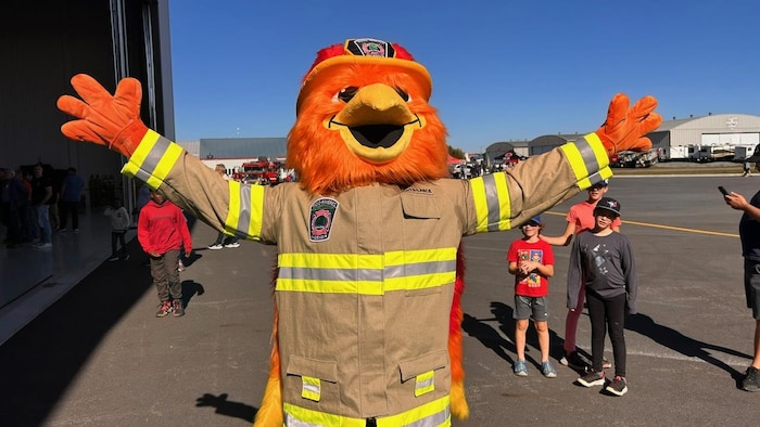 Une mascotte représentant un phénix vêtu d’un uniforme de pompier lève les deux bras dans les airs, dehors.