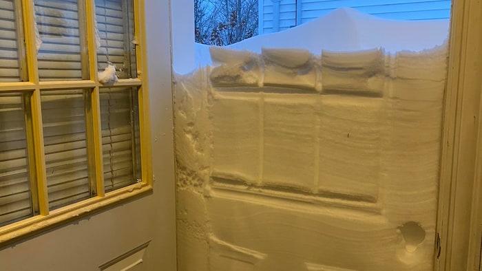 Une porte ouverte laisse voir un demi mur de neige qui bloque l'accès et empêche de sortir d'une maison.