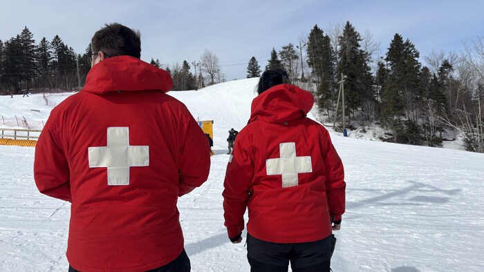Vue de dos, deux patrouilleurs de ski portent des manteaux rouges arborant une grande croix blanche centrale. Ils se tiennent debout sur une piste enneigée, faisant face à une pente de ski bordée d'arbres sous un ciel dégagé.