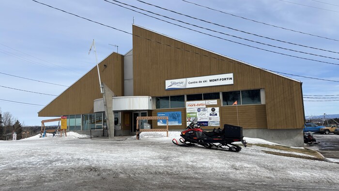 Cette photo montre le pavillon principal du centre de ski Mont-Fortin à Saguenay. Le bâtiment brun à l'architecture angulaire surplombe un terrain glacé où stationne une motoneige noire. Des fils électriques traversent le ciel clair en arrière-plan.