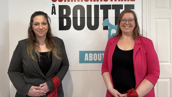 Mylène Beaulieu et Mélanie Martel portent le tissu rouge sur leurs bras devant une affiche du mouvement « Le Communautaire à boutte! ».