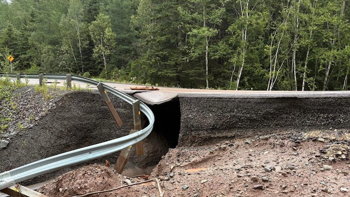 Une route asphaltée traversant une forêt est totalement effondrée et coupée en deux par une profonde crevasse causée par des inondations, laissant voir une glissière de sécurité déformée suspendue au-dessus du vide