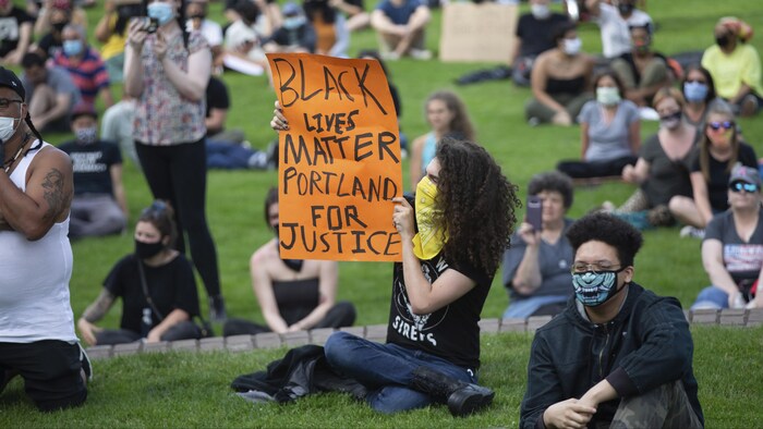 Des manifestants assis sur la pelouse dans un parc, une femme tient une affiche sur laquelle elle a écrit « Black lives matter, Portland for justice ».