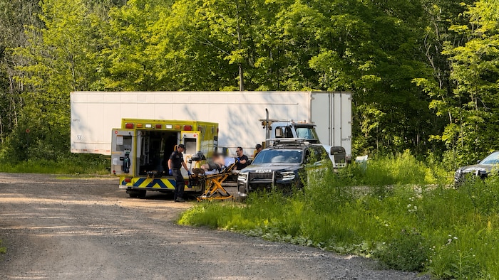 Un homme, dont le visage est flou, est attaché sur une civière à côté d'une ambulance et d'une voiture de police. 