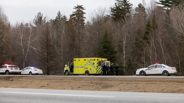 Deux véhicules de police et une ambulance devant un boisé, le long d'une autoroute.