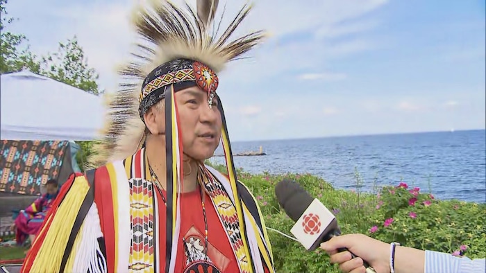 Un homme vêtu d'une tenue traditionnelle autochtone avec une coiffe à plumes est interviewé à l'extérieur. Un microphone de Radio-Canada est tendu vers lui. À l'arrière-plan, on aperçoit un plan d'eau, de la végétation et un ciel bleu.