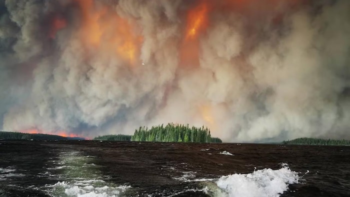 Un incendie fait rage au sein de la Première Nation de Collins tandis qu’une personne évacuée s’enfuit en traversant le lac Collins, le 14 juillet 2026.