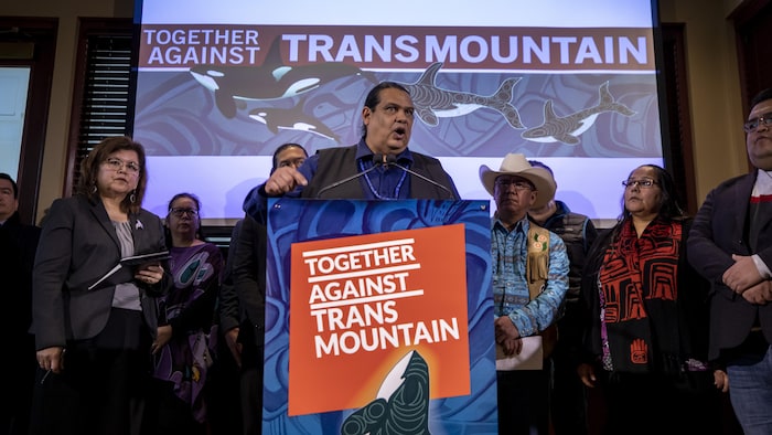 Un groupe de chefs autochtones debout derrière un podium sur lequel est écrit en anglais « Ensemble contre Trans Mountain ».