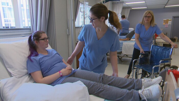 Une femme attend avec un fauteuil roulant, alors qu'une autre aide une patiente fictive à se lever de son lit.