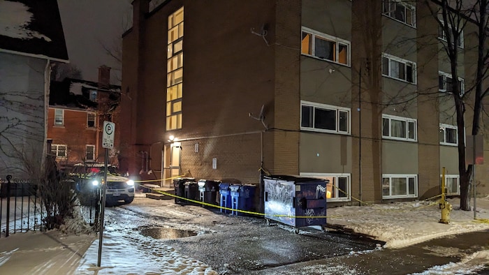 Du ruban jaune encercle des poubelles et un conteneur à déchets devant un immeuble de logements.