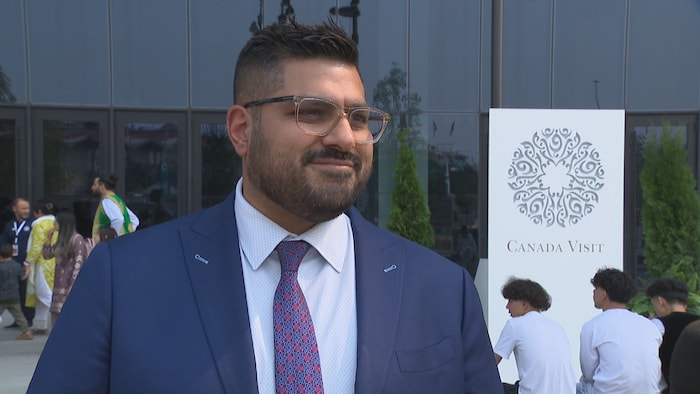 Un homme vêtu d'un complet bleu marine, d'une chemise claire et d'une cravate à motifs, porte des lunettes à monture transparente et sourit légèrement à l'extérieur, devant un bâtiment vitré et une pancarte blanche indiquant « CANADA VISIT ».