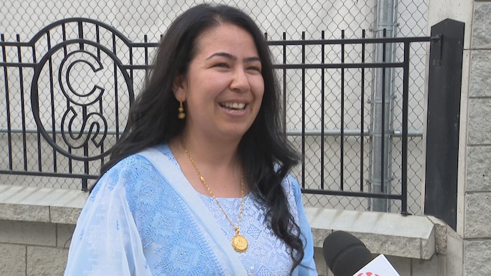 Une femme sourit lors d'une entrevue au microphone de Radio-Canada, vêtue d'un vêtement bleu clair brodé et portant un collier en or, debout à l'extérieur devant une clôture métallique.