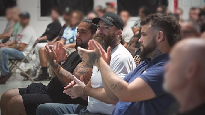Plusieurs hommes assis applaudissent en regardant vers l'avant. Au premier plan, trois hommes non floutés, dont un avec des tatouages sur les bras, applaudissent attentivement. En arrière-plan, les visages des autres détenus sont brouillés.