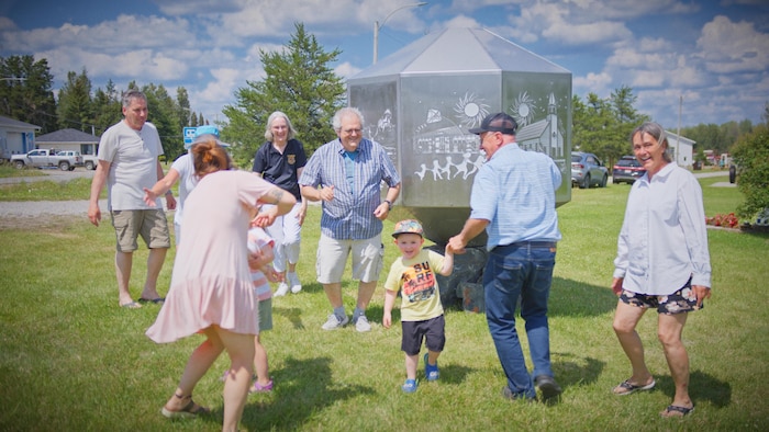 Photo thématique sur laquelle des gens de différentes générations dansent sur la pelouse, devant une sculpture.