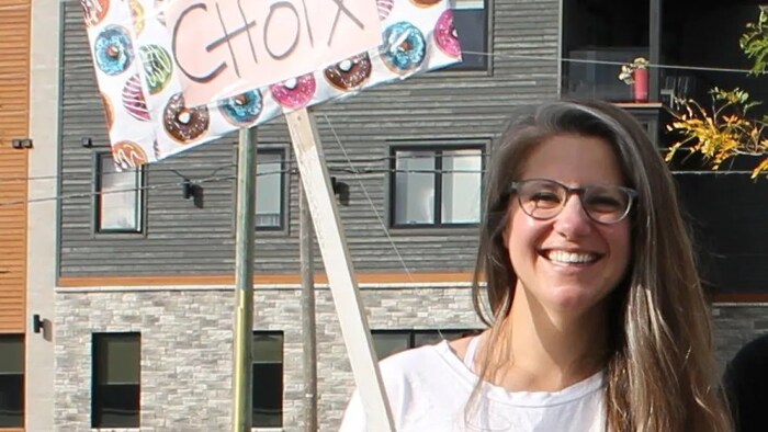 Une femme aux cheveux longs portant des lunettes. Elle se tient devant une maison. Elle tient une pancarte sur laquelle est écrit ''pro-choix''.