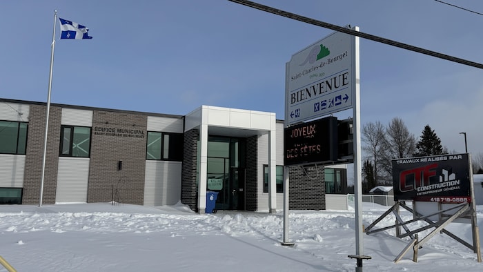 Cette photo montre l'édifice municipal de Saint-Charles-de-Bourget sous la neige. Un drapeau du Québec flotte à gauche, tandis qu'un grand panneau de bienvenue et une affiche de construction occupent le premier plan à droite sous un ciel clair.