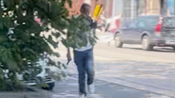 Damien Hudson marche de dos dans une rue, une arme à feu à la main droite.