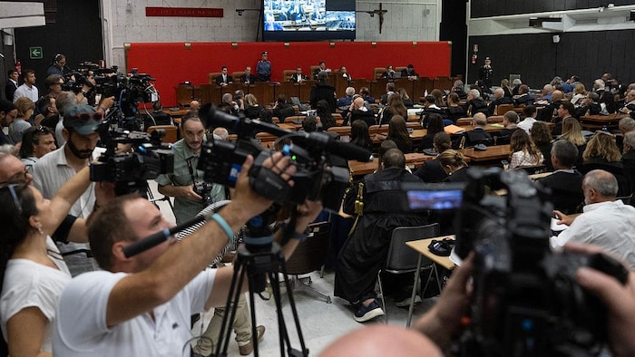 Des juges assis devant une grande salle d'audience remplie de journalistes, photographes et caméramans.