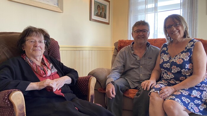 Une dame assise dans un fauteuil et un couple assis sur une causeuse posent pour la caméra en souriant.