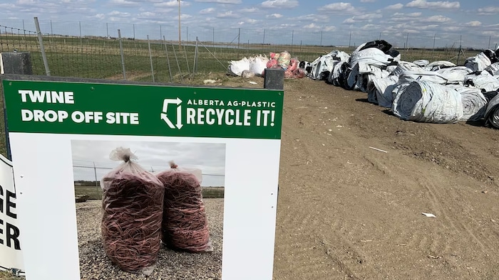 « AlbertaAg-Plastic, Recycle it! » Des ballots de sacs-silos à grain et de ficelles d'enrubannage sont sur le sol près de l'affiche.