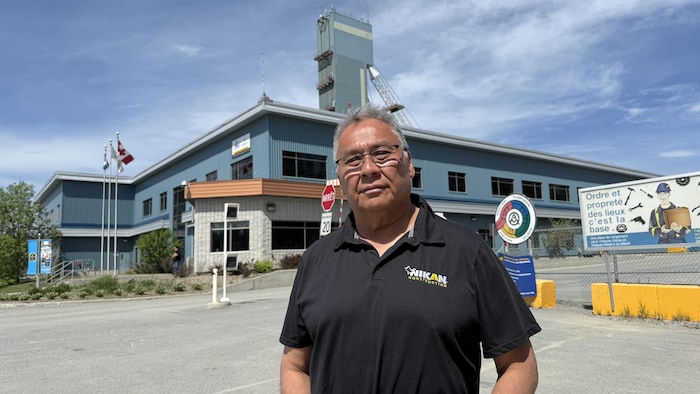 John Mowatt pose pour la caméra devant un bâtiment de deux étages et un chevalement de mine.