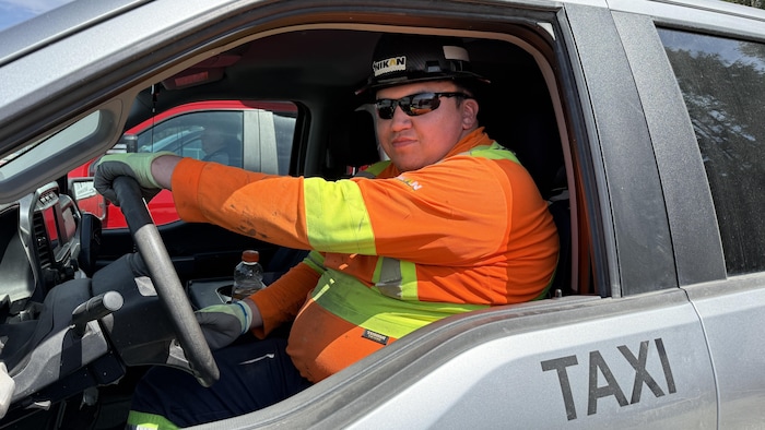 Taylor Mapachee, assis au volant d'un véhicule taxi, regarde le caméra munis de ses lunettes de soleil.
