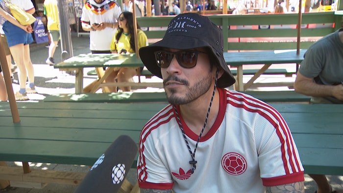 Un homme avec un maillot de sport, des lunettes et un chapeau parle à un micro.
