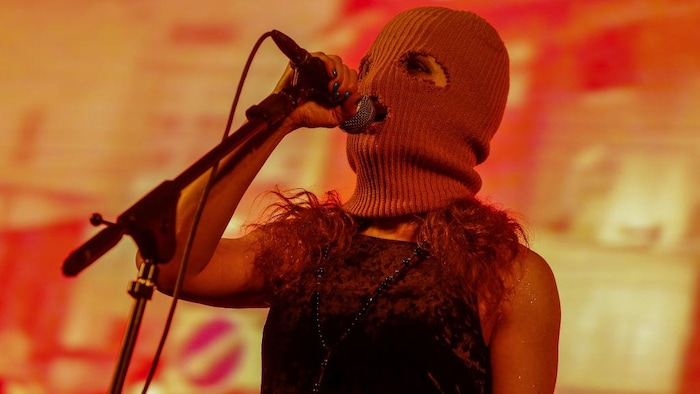 A woman in a balaclava sings into a microphone.
