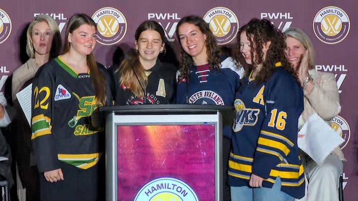 Ivy Johnson, Elyse Tidridge, Brianne Graves et Emma McLeod debout devant un podium, souriantes et vêtues de maillots de hockey.