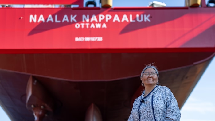 Une femme, les deux mains dans les poches, le regarde posé fièrement sur l'horizon, avec derrière elle un haut navire rouge décoré du nom Naalak Nappaaluk en lettres blanches.