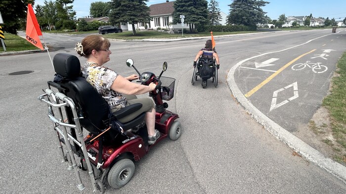 Une femme en quadriporteur et une autre en fauteuil roulant contournent une piste cyclable.