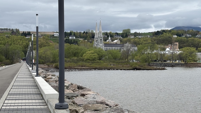 Sous un ciel nuageux s'élèvent les clochers de la basilique Sainte-Anne-de-Beaupré en arrière-plan. À l'avant-plan, une bande de béton et de pavé formant un quai piétonnier protégée des vagues par un enrochement.