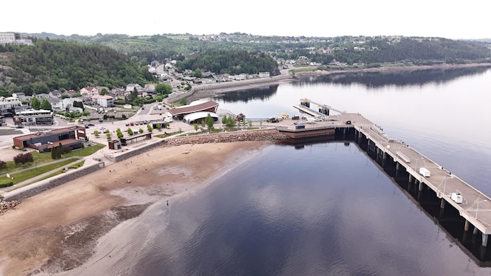 Un long quai s'avance dans l'eau, à côté d'une plage de sable, de bâtiments côtiers et d'une colline habitée.