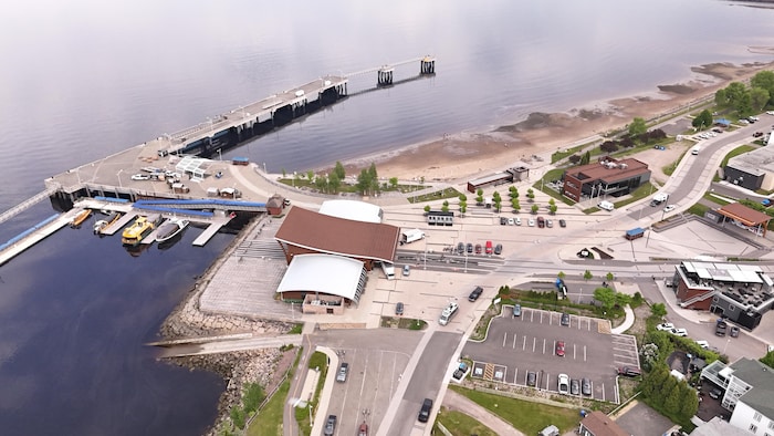 Un grand quai et des bateaux bordent une place publique dotée d’un pavillon moderne, de stationnements et d’une plage.
