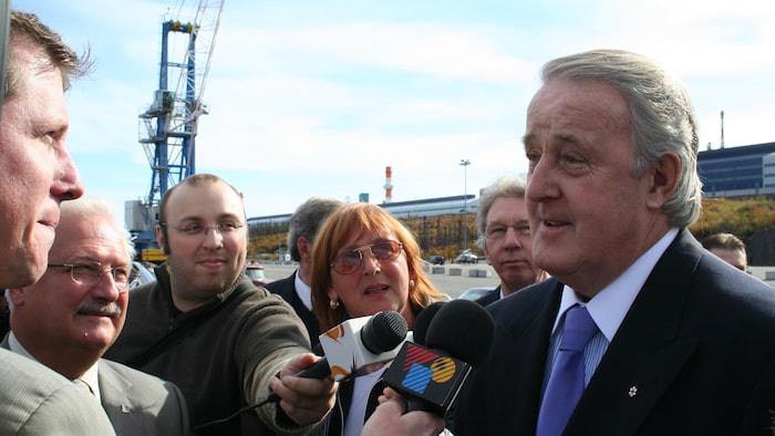 Brian Mulroney entouré par des journalistes, micros en main.