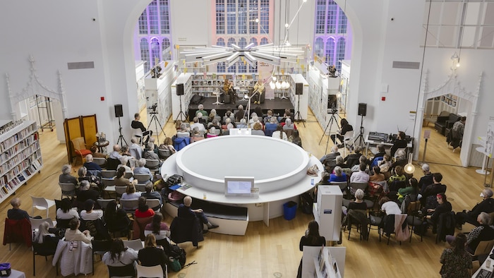 Un public assis écoute une présentation donnée sur une scène au fond d'une grande bibliothèque lumineuse.