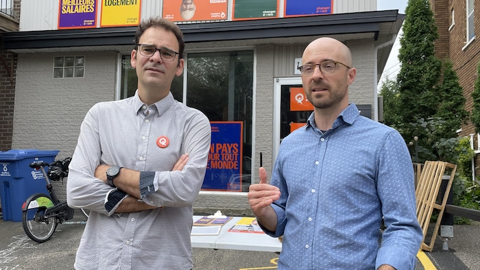 Deux hommes se tiennent devant un immeuble où l'on peut voir des affiches de Québec solidaire.