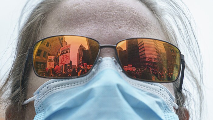 Une femme en gros plan porte un couvre-visage et des lunettes de soleil dans lesquelles on voit le reflet d'une manifestation, avec des pancartes brandies et des gratte-ciel.