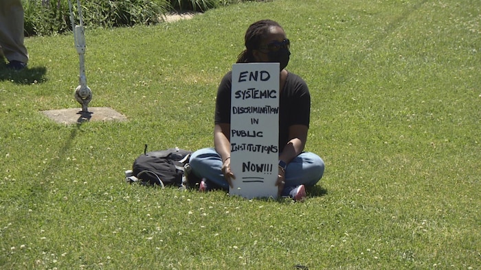 Une manifestante tient une pancarte où il est écrit qu'il faut faire cesser la discrimination systémique au sein des institutions publiques.