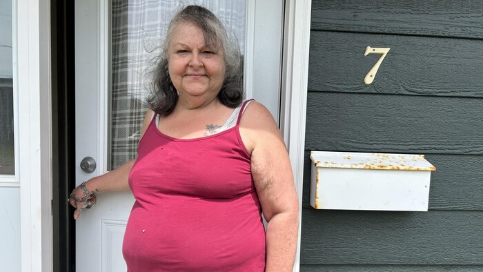 Une femme sourit à la caméra, à l'entrée de son logement, tenant la poignée de sa porte.