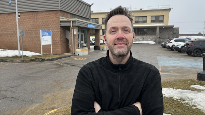 Le Dr. Aubert regarde la caméra les bras croisés devant le centre multiservices.