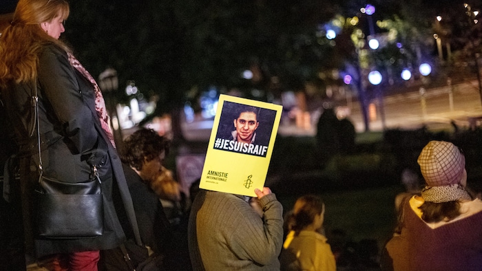 Plusieurs personnes rassemblées dehors, la nuit. L'une d'elles tient une affiche qui cache sa tête.