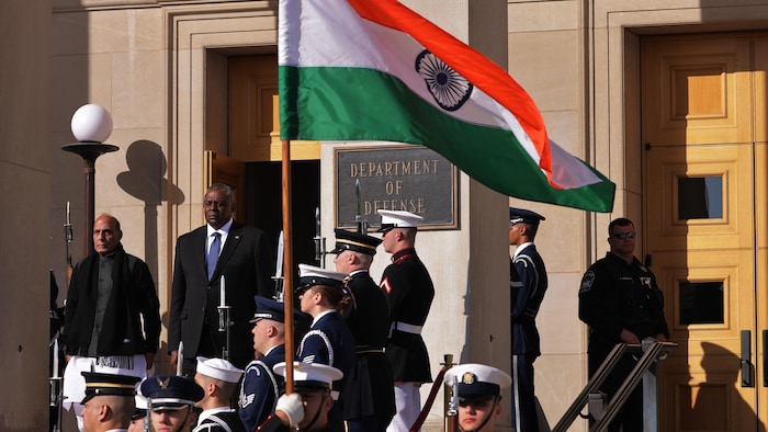 Rajnath Singh et Lloyd Austin se tiennent debout à l'extérieur devant un cortège militaire. Un drapeau indien flotte.