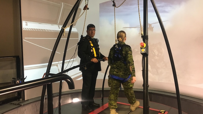 La capitaine Anna Harpe marche sur un tapis roulant alors que l'image d'un hélicoptère atterrissant dans un nuage de poussière est projetée devant elle. Le colonel Jetly est debout près d'elle et lui parle.