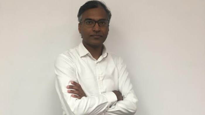Ram Rammaya pose pour la photo, il a les bras croisés, il porte une chemise blanche et un pantalon noir avec une ceinture noire. Il a la peau foncée, il porte des lunettes, il a les cheveux noirs avec un peu de blanc sur les bords.