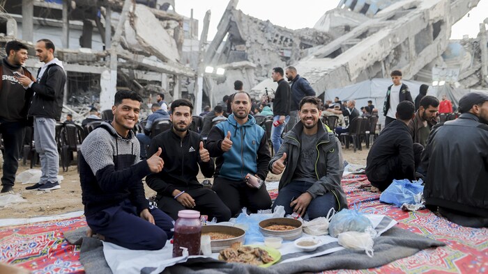 Des hommes partagent un repas sur un tapis étendu entre les décombres, le 15 mars 2025. Derrière eux, une grande tour s'est effondré.
