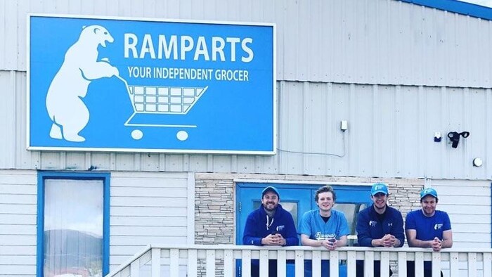 Quatre hommes se tiennent sur une rambarde, en dessous d'un logo d'épicerie.