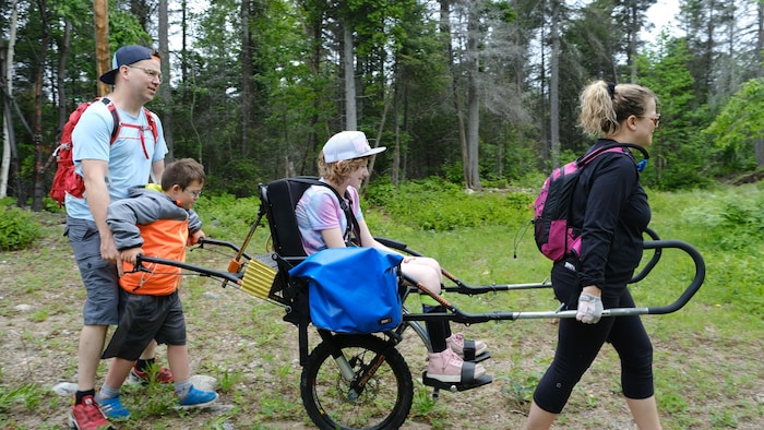 Une personne dans un fauteuil roulant adapté est transportée par deux autres personnes dans un sentier en forêt.